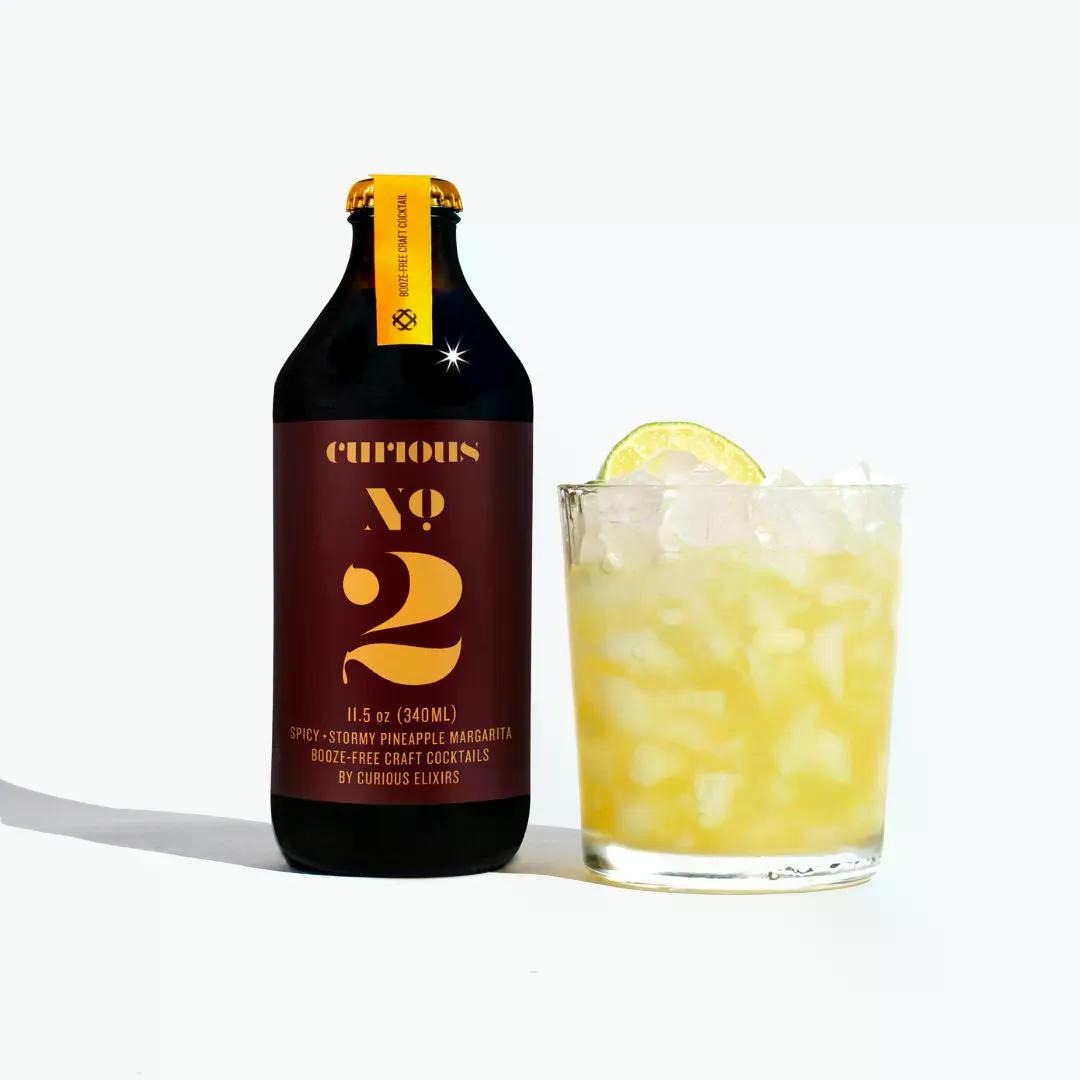 Bottle of Curious Elixir no. 2 Spicy Pineapple Margarita next to a glass filled with ice and a lemon garnish