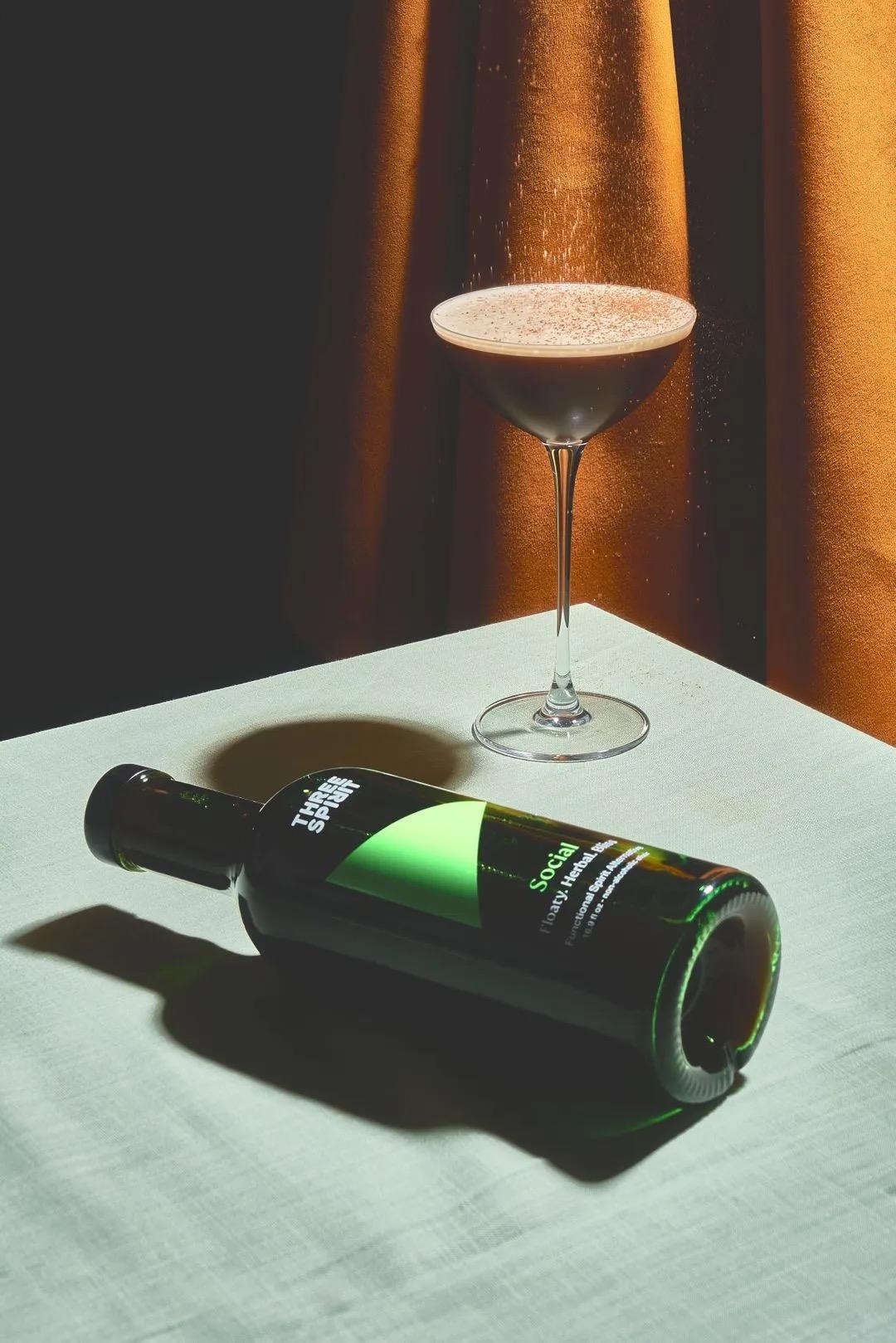 Bottle of Three Spirits Social Elixir next to a filled cocktail glass on a white table with warm lighting