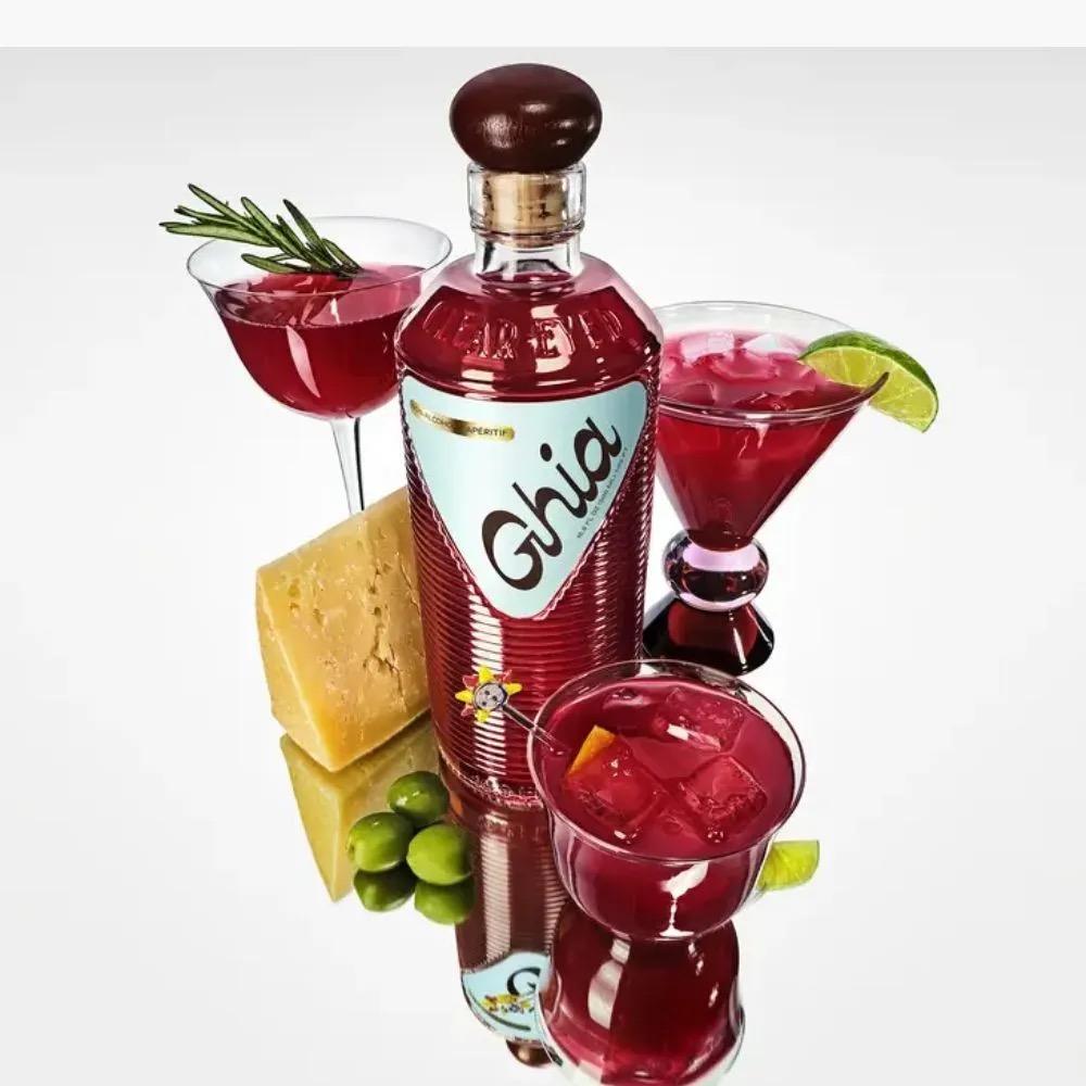 Bottle of Ghia Original Apéritif with three red cocktails, cheese wedge, and green olives on reflective surface.