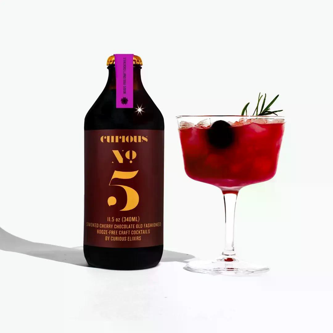 Curious Elixer No 5 bottle with a red non-alcoholic botanical cocktail garnished with a cherry and rosemary on a white background