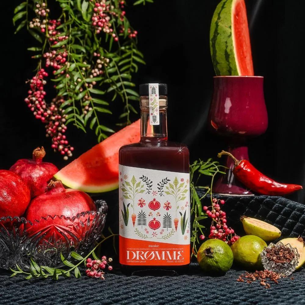 Bottle of Drømme Awake botanical blend surrounded by pomegranates, watermelon, chili pepper, and fresh herbs on black background