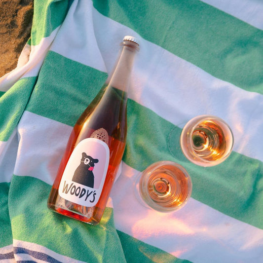 Bottle of Woody's Sparkling Rosé and two glasses on green and white striped towel outdoors in sunlight