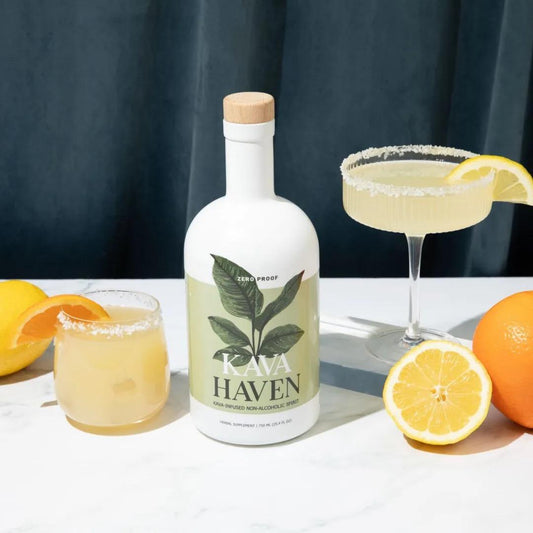 Bottle of Kava Haven non-alcoholic spirit with two lemon-ginger cocktails and fresh citrus on a white table