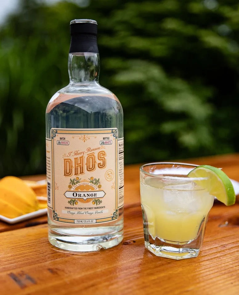 Transparent bottle of Dhōs Orange non-alcoholic liqueur next to a citrus cocktail with lime garnish on wooden surface