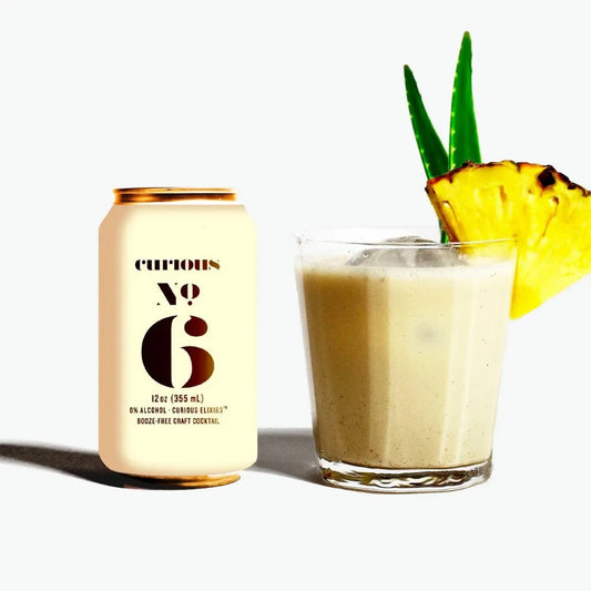Curious Elixir No 6 Piña Colada can with creamy vegan cocktail in glass garnished with pineapple slice and green leaves