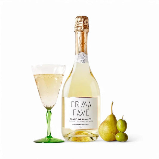 Bottle of Prima Pavé Blanc de Blancs alcohol-free sparkling wine next to a glass and fresh fruit on white background