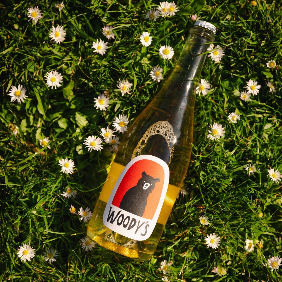 Bottle of Woody's sparkling non-alcoholic Blanc de Blanc on grass with white daisies around it